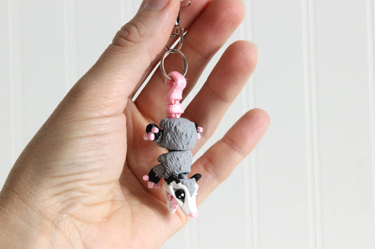 Hand holding the 3D-printed gray-and-white opossum keychain to show its size, colors, and silver swivel clip.