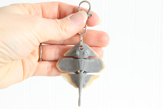 Hand holding the 3D-printed gray stingray keychain to show its size, colors, and silver swivel clip.