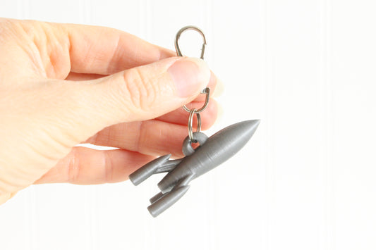 Hand holding the 3D-printed rocket keychain to show its size, colors, and silver swivel clip.
