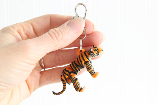 Hand holding the 3D-printed orange, black, and white tiger keychain to show its size, colors, and silver swivel clip.