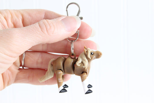 Hand holding the 3D-printed brown horse with a blonde mane keychain to show its size, colors, and silver swivel clip.