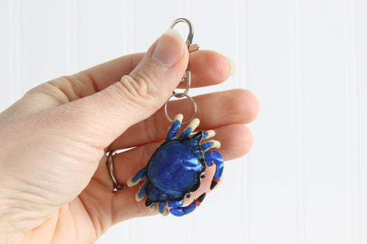 Hand holding the 3D-printed blue crab keychain to show its size, colors, and silver swivel clip.