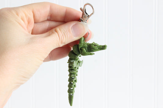 Hand holding a green alligator keychain, showing its segmented body and silver swivel quick-clip.