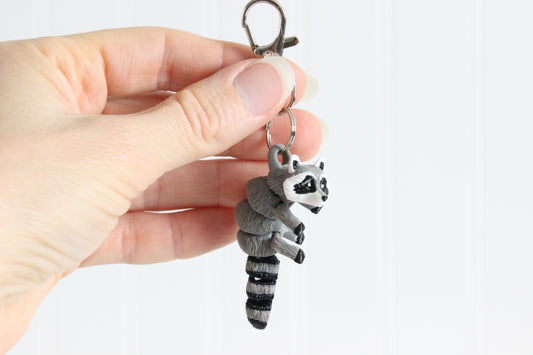 Hand holding the 3D-printed gray-and-black raccoon keychain to show its size, colors, and silver swivel clip.