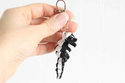 Hand holding the 3D-printed black-and-white skunk keychain to show its size, colors, and silver swivel clip.