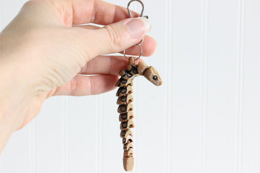Hand holding the 3D-printed tan-and-brown rattlesnake keychain to show its size, colors, and silver swivel clip.