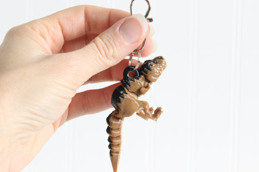 Hand holding the 3D-printed T. rex dinosaur keychain to show its size, colors, and silver swivel clip.