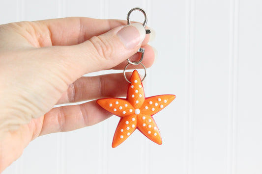 Hand holding the 3D-printed orange starfish keychain to show its size, colors, and silver swivel clip.