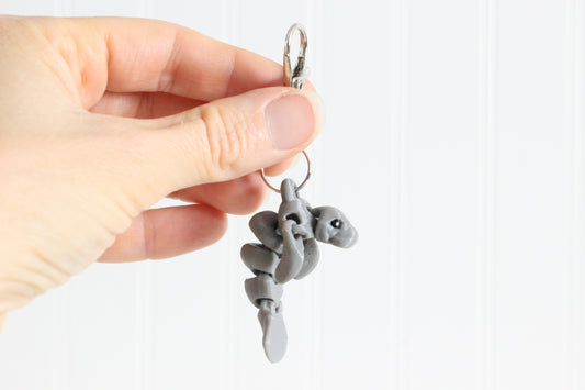 Hand holding the 3D-printed gray manatee keychain to show its size, colors, and silver swivel clip.