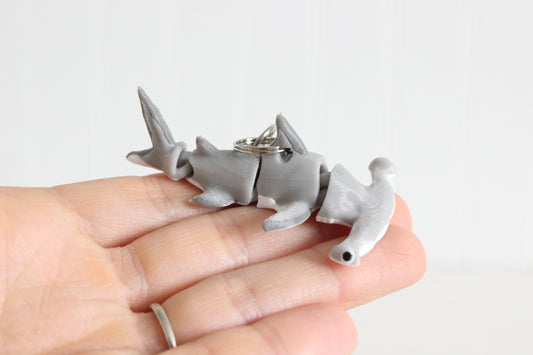 Hand holding the 3D-printed gray hammerhead shark keychain to show its size, colors, and silver swivel clip.