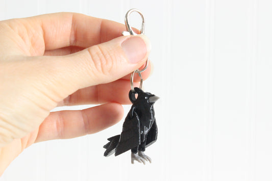 Hand holding the 3D-printed black raven keychain to show its size, colors, and silver swivel clip.