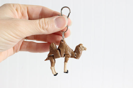 Hand holding the articulated camel keychain to show its size, textured fur, and cream-colored legs.
