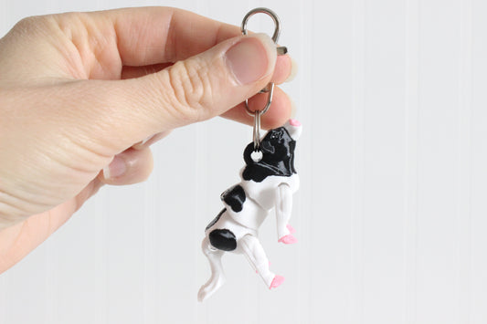 Hand holding the 3D-printed black-and-white cow keychain to show its size, colors, and silver swivel clip.