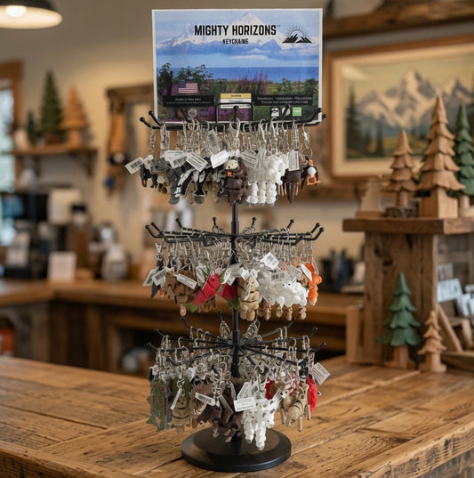 A multi-tier display stand holding assorted adorable keychains, set on a rustic wooden counter inside a cozy shop. The top card shows a scenic display card reading 'MIGHTY HORIZONS'